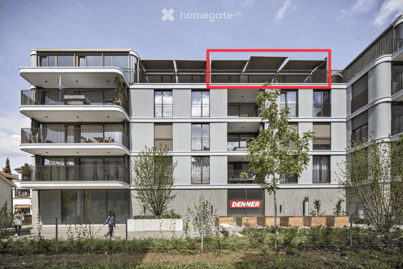 Modern multi-story residential building with balconies, large windows, and contemporary design. Features include a rooftop terrace with outdoor seating and a fenced garden area at the base.