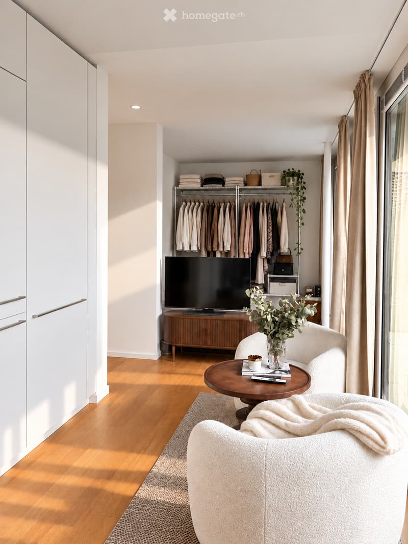 Modern living room with a white sofa, wooden coffee table, flat-screen TV, and a wardrobe with hanging clothes.
