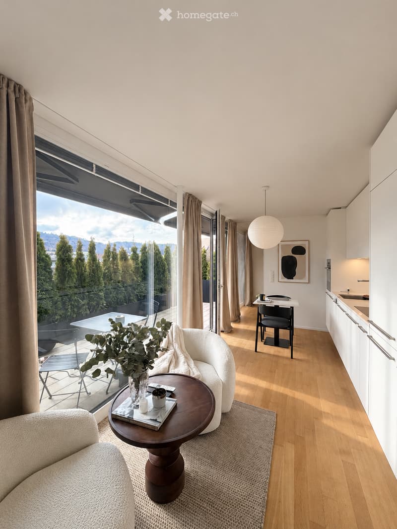 Modern living room with large windows offering a scenic view, featuring a white sofa, round coffee table, dining area with chairs, and a small kitchen area with cabinets.