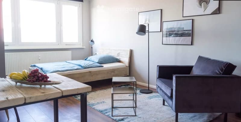 A modern, well-lit room with a bed, sofa, small table, and floor lamp. The room features a wooden floor, large windows, and a decorative bowl of fruit on the table.