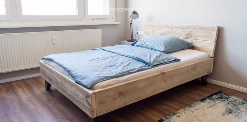 Cozy bedroom with a wooden bed, blue bedding, a nightstand with a lamp, and a patterned rug on the floor.