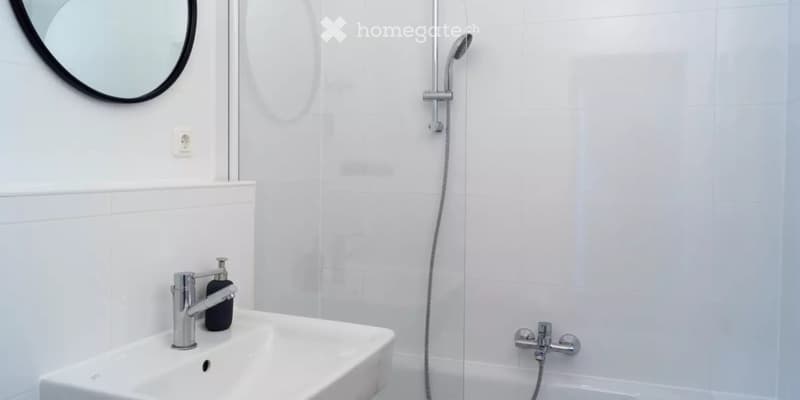 Modern bathroom with white tiles, a sink with a chrome faucet, a showerhead, and a round mirror on the wall.