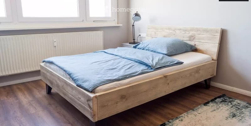 A modern bedroom with a wooden bed, blue bedding, a nightstand with a lamp, and a radiator under the windows.