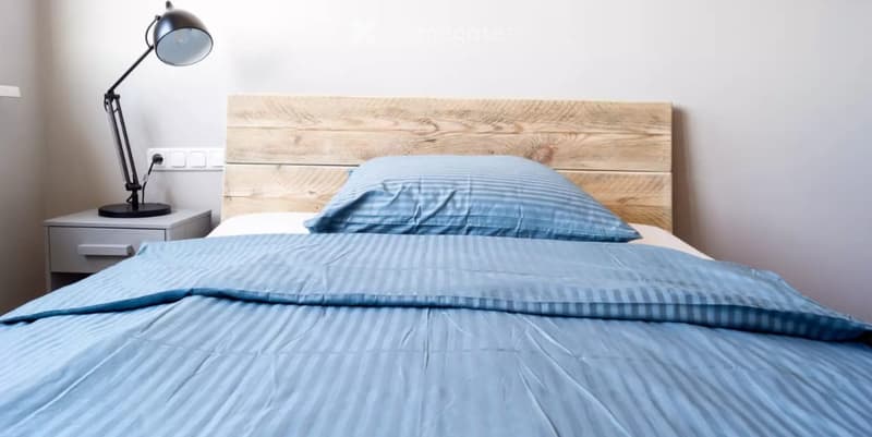A modern bedroom with a wooden headboard, a neatly made bed with blue bedding, a nightstand, and a lamp.