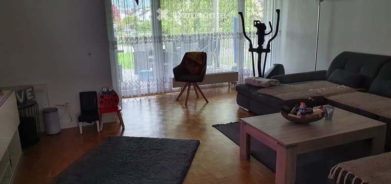 Living room with wooden flooring, large window with sheer curtains, sofa, coffee table, exercise bike, and small decorative items.