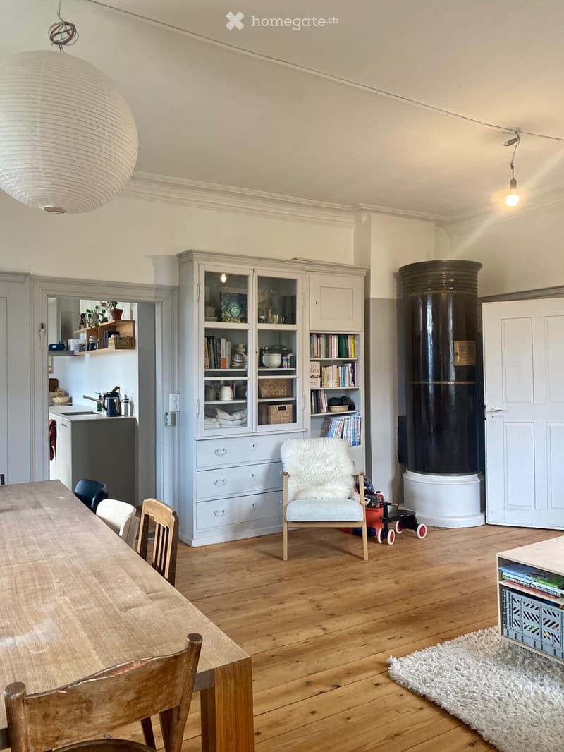 A well-lit living space with wooden flooring, a large wooden dining table with chairs, a cabinet with glass doors, a futon chair, a small kitchen area with a sink and refrigerator, and a toy car on the floor.