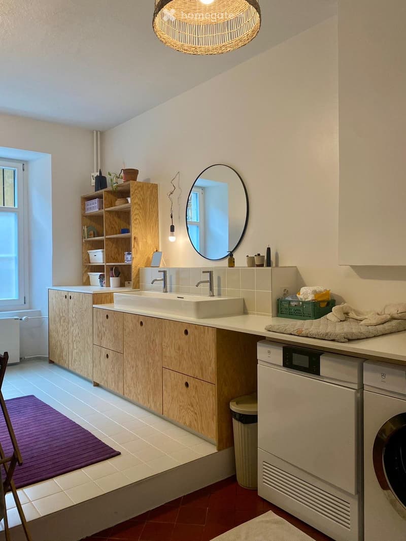 Modern bathroom with a double sink, wooden cabinets, a large round mirror, a shelf with decorative items, a washing machine, and a drying rack with towels.