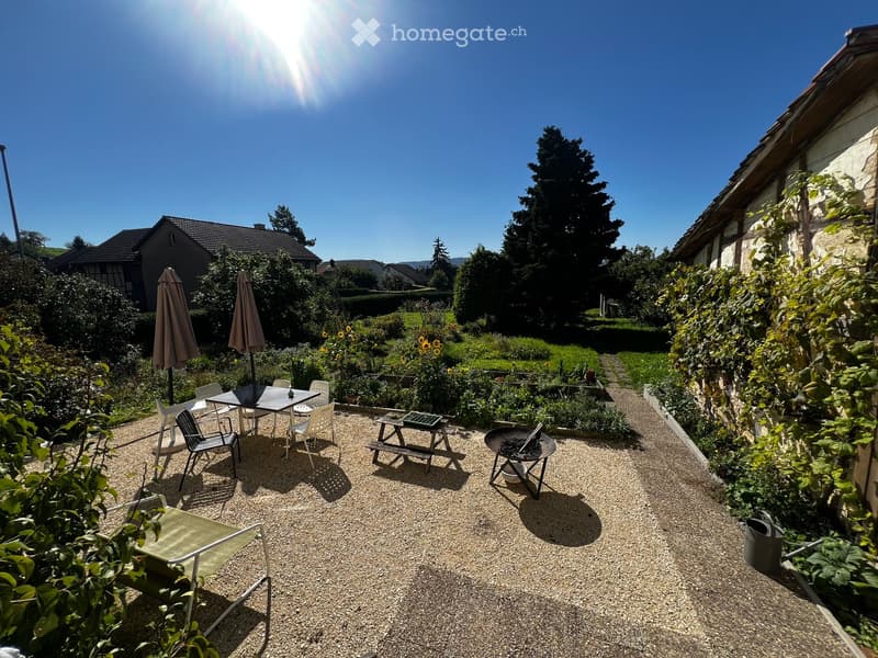 Garden area with gravel ground, outdoor furniture including a table, chairs, umbrellas, a picnic table, and a fire pit, surrounded by greenery and flowers.