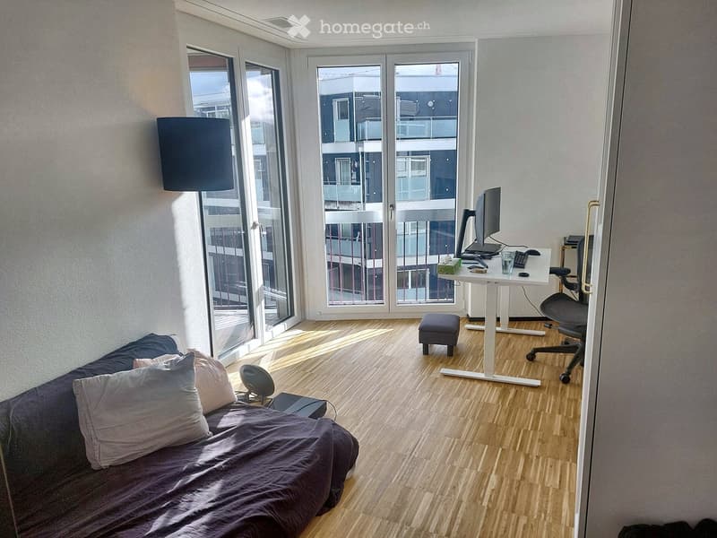 A modern, multi-functional room with a sofa bed, a desk setup, and large windows offering a view of the building exterior.