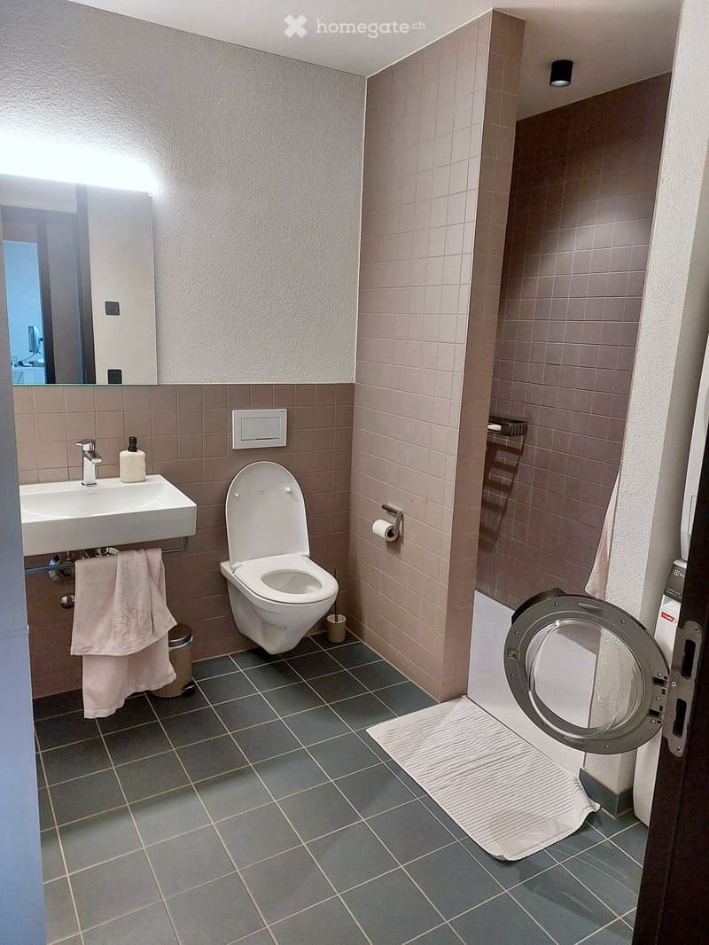 Modern bathroom with a white sink, toilet, towel, and mirror. Grey tiled floor and beige tiled walls.
