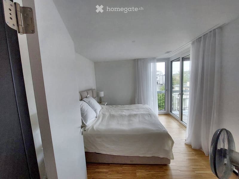 Modern bedroom with a large bed, nightstand, lamp, and a fan near the window. The room features wooden flooring and large windows with white curtains, offering a view of the balcony and outdoor area.