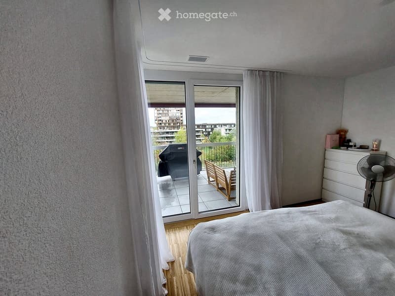 Bedroom with a bed, a fan, a dresser, and a table, with a sliding glass door leading to a balcony.