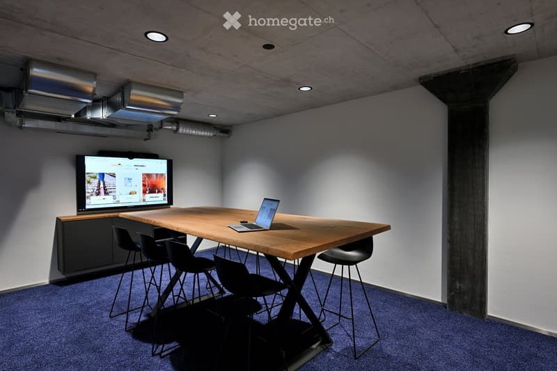An empty office room with a rectangular wooden table with a laptop on it, surrounded by four high black chairs, a flatscreen tv mounted on a cabinet, and a dark blue carpet.