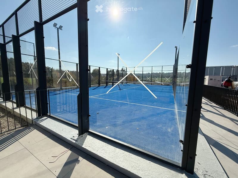 A modern, enclosed tennis court with a blue playing surface, surrounded by glass walls and metal fencing, under a clear blue sky.