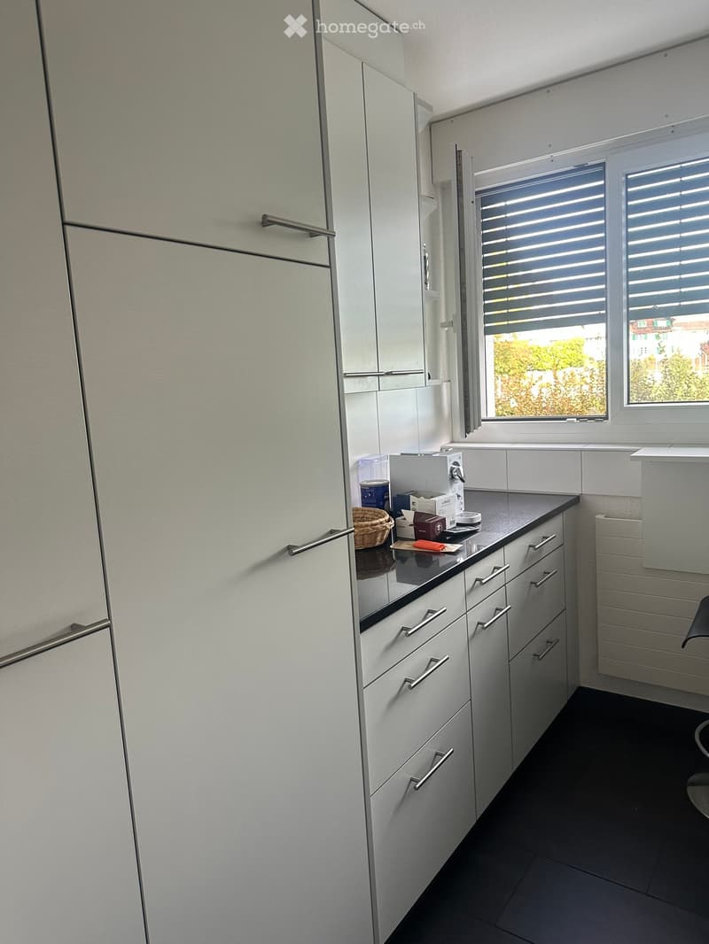 Modern kitchen with white cabinets, dark countertops, and a window with blinds. Visible items include a small basket and a few containers on the counter.
