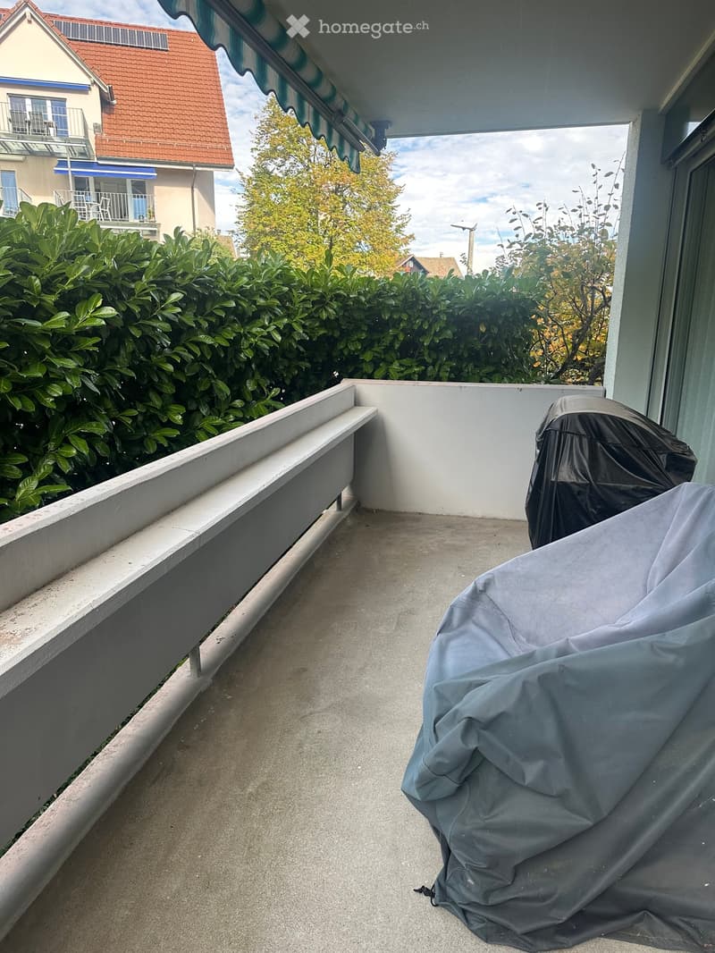 Balcony with covered furniture, white railing, and view of neighboring building and greenery.