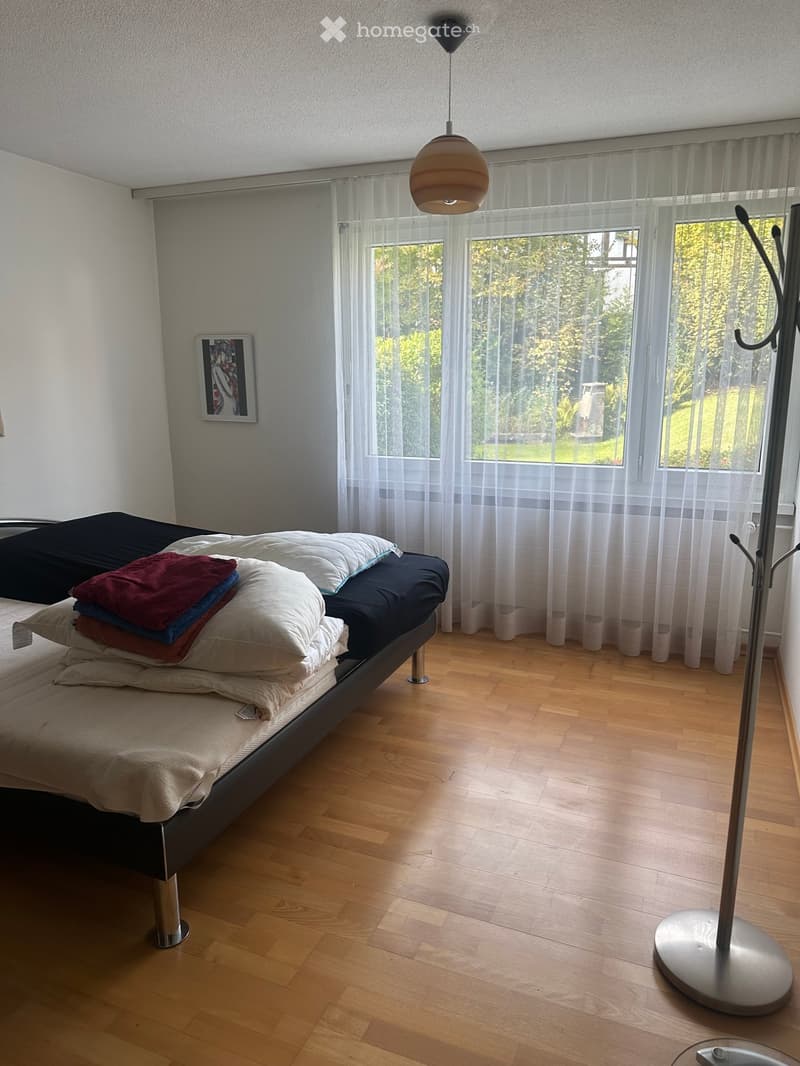 Bright bedroom with wooden flooring, large window with sheer curtains, bed with multiple pillows and blankets, picture frame on wall, and a floor lamp.