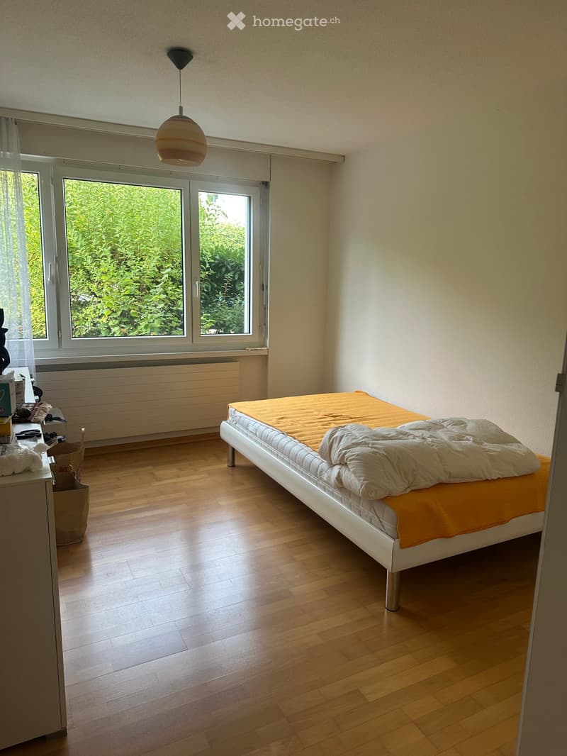 Bright bedroom with wooden flooring, large windows, a white bed with orange bedding, a nightstand, and a hanging lamp.