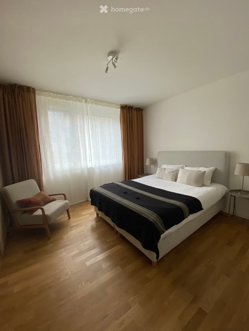Modern bedroom with wooden flooring, large bed with white headboard and black and white bedding, a white armchair with a red pillow, and two table lamps on nightstands.