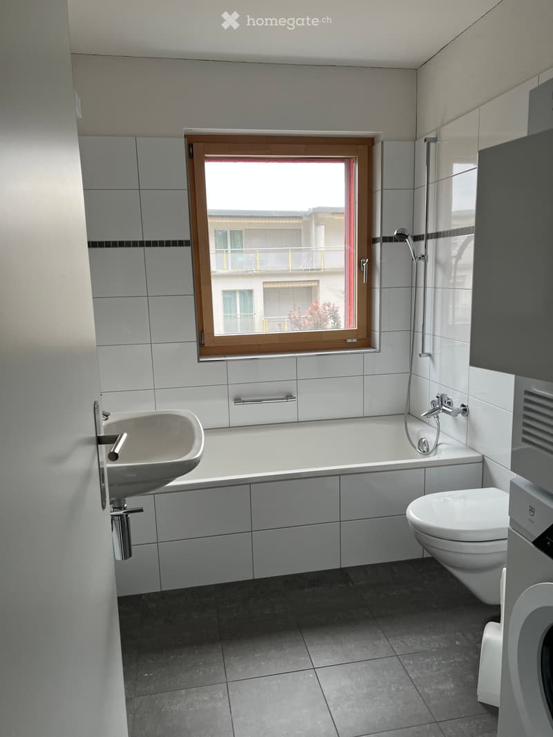 Modern bathroom with a bathtub, toilet, sink, and showerhead. Features a large window with wooden frame and a view of a neighboring building.
