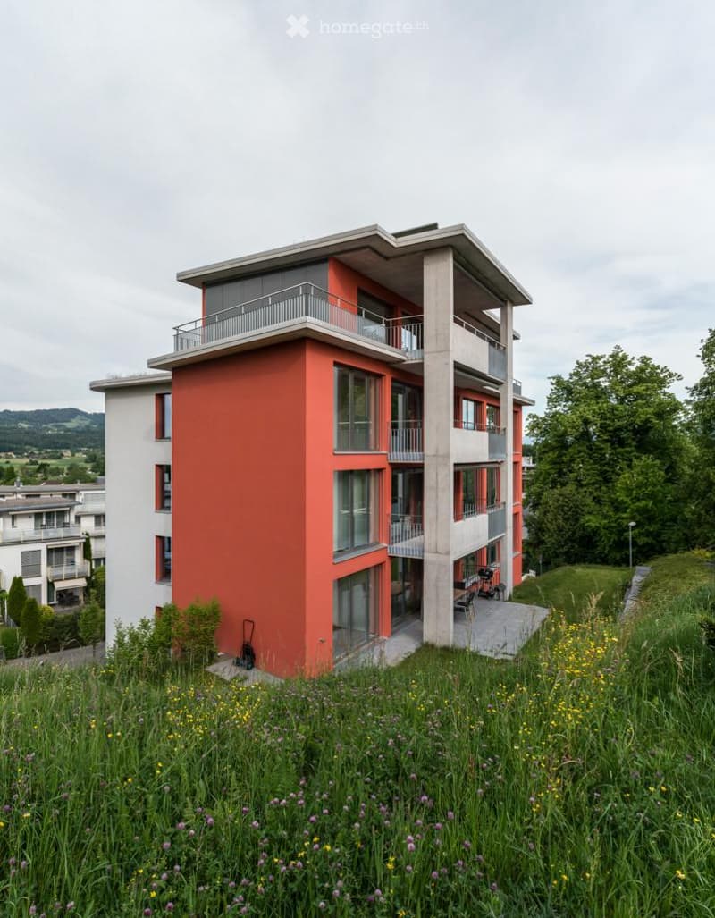 Modern multi-story building with red and white facade, large windows, balconies, and a raised ground floor.