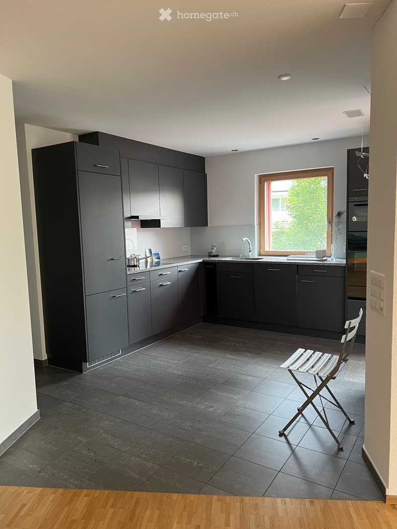 Modern kitchen with grey cabinets, dark tiled floor, and a single white folding chair near the window.