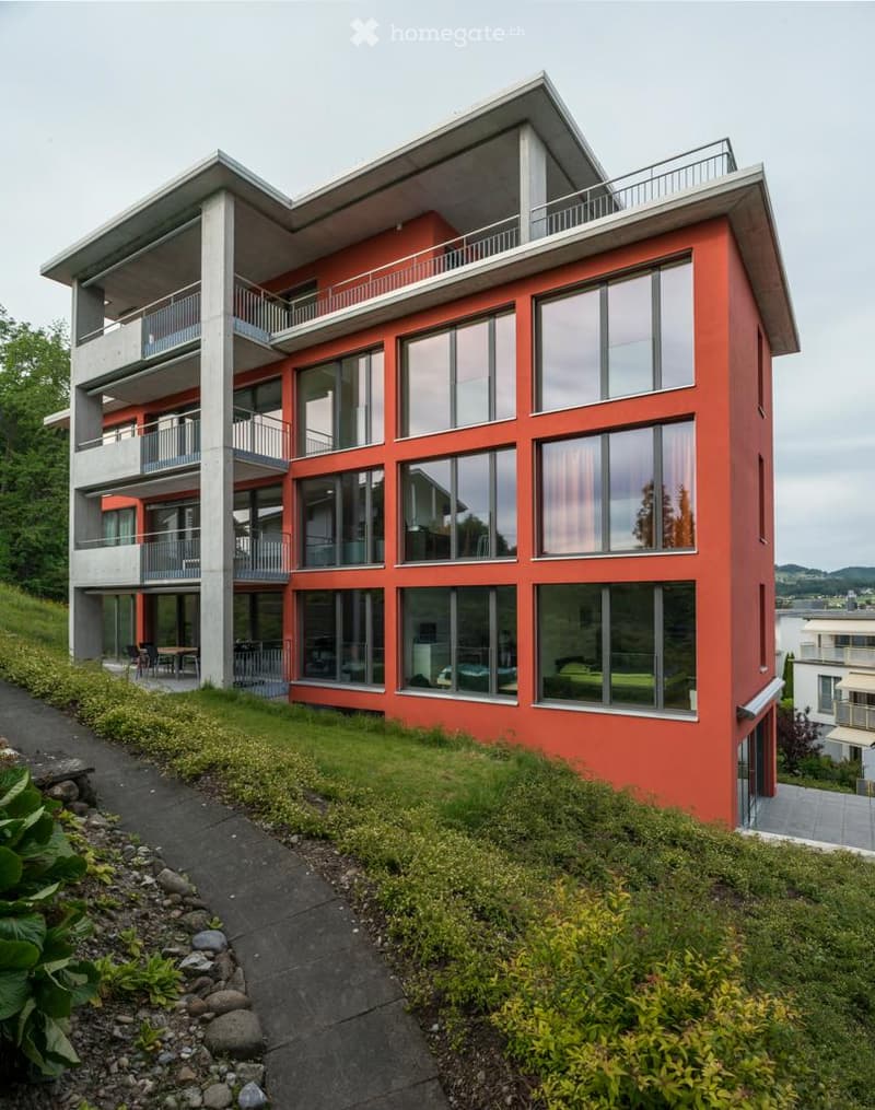 Modern, multi-story building with red-orange facade and large windows, featuring balconies and a raised ground floor.