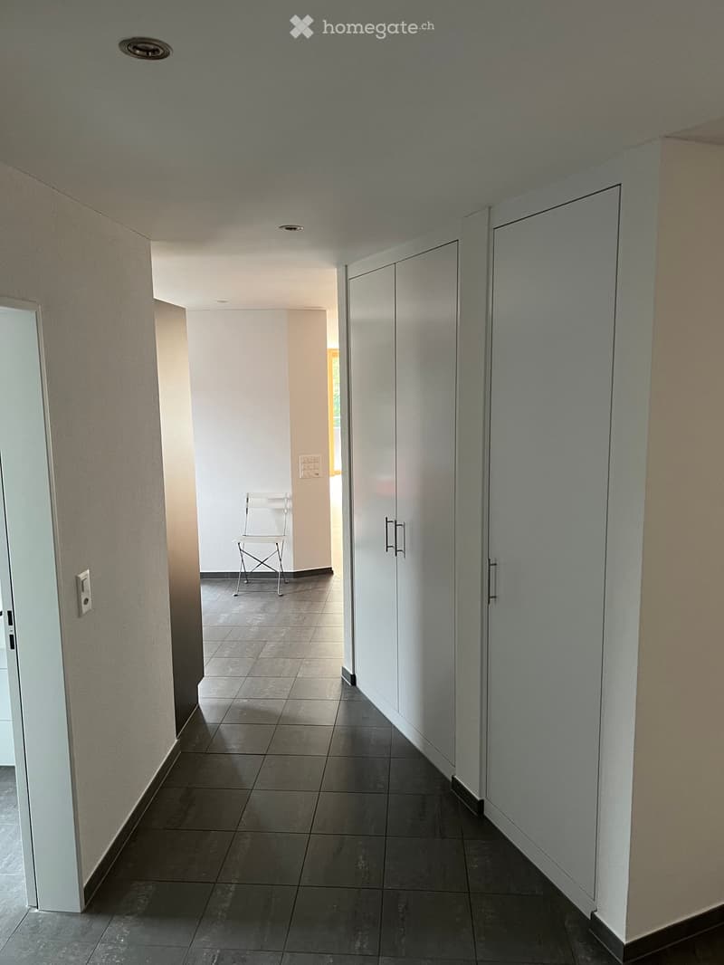 Empty hallway with tiled floor, white walls, built-in cabinets, and a single chair in the background.