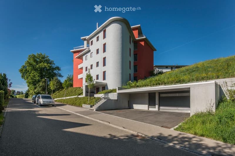 Modern multi-story building with a curved white facade and red accents, featuring a covered garage and parking area.