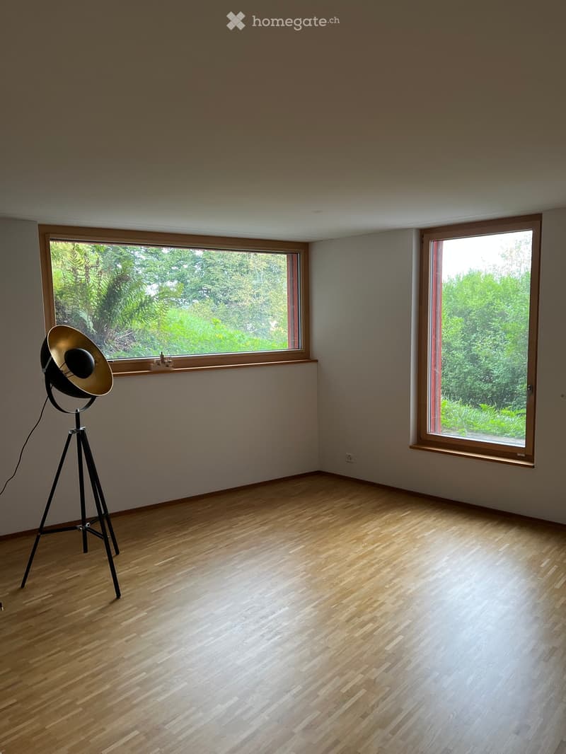 Empty room with wooden flooring, large windows, and a single tripod-mounted lamp. No furniture or equipment present.