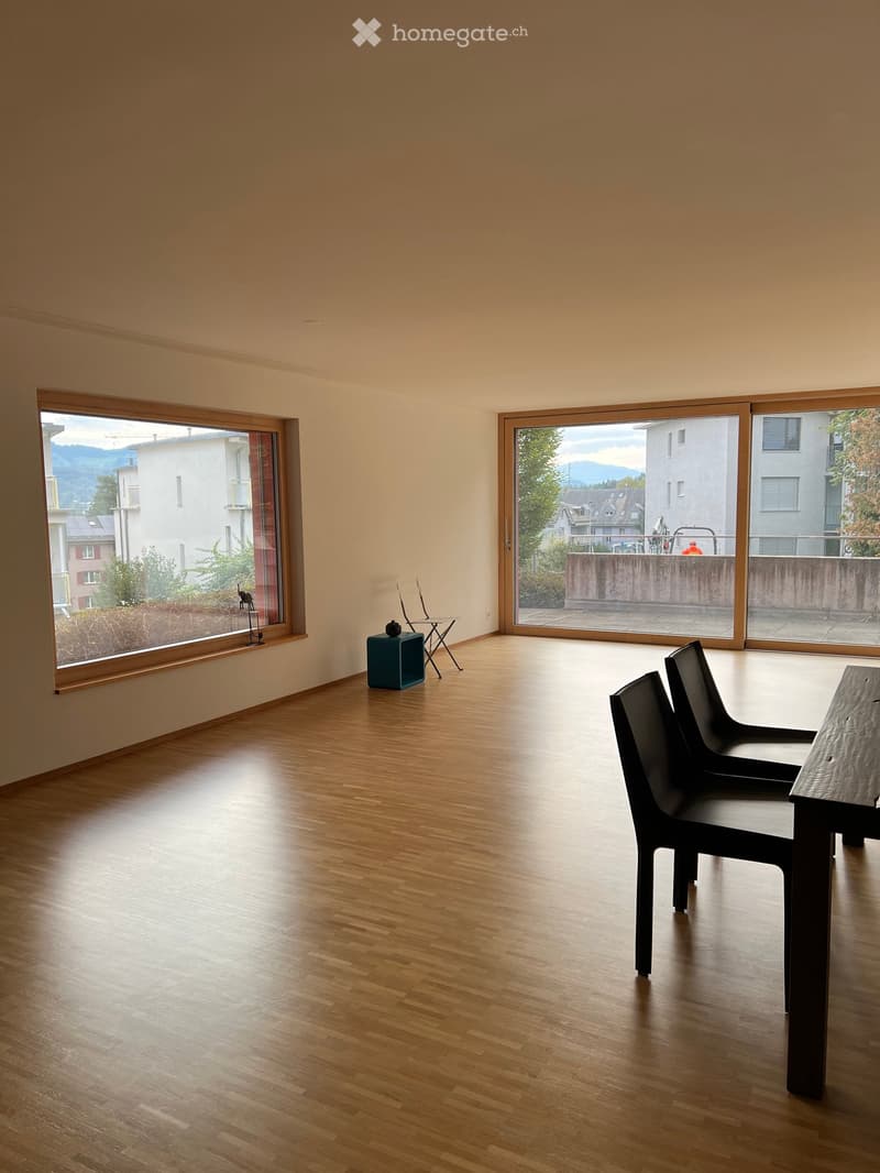 Bright, unfurnished room with wooden flooring, large windows, and a few modern chairs and a small table.