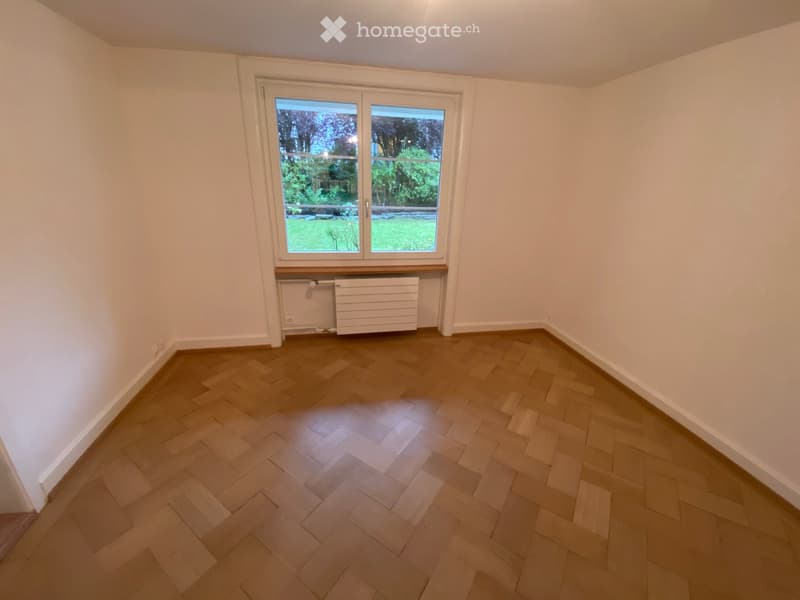 Empty room with wooden herringbone flooring, white walls, large window with view of greenery, and radiator below the window.