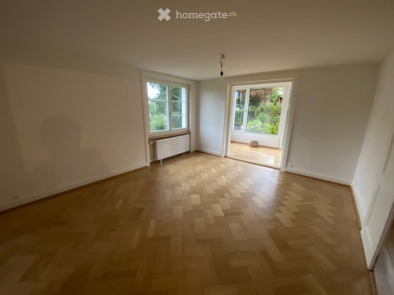 Empty room with wooden herringbone flooring, white walls, large windows, and a radiator. A door leads to a balcony or outdoor area.