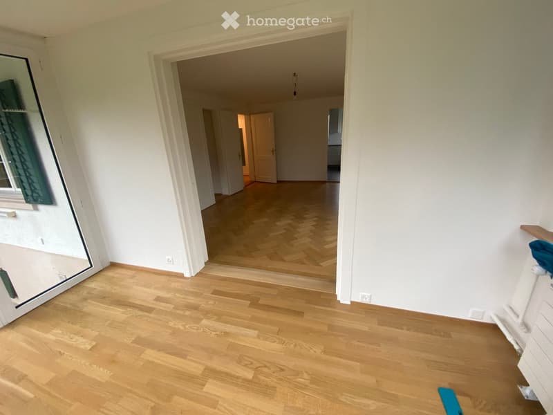 Empty room with light wooden flooring, white walls, and a large mirror on one wall. The room has a doorway leading to another space with similar flooring.