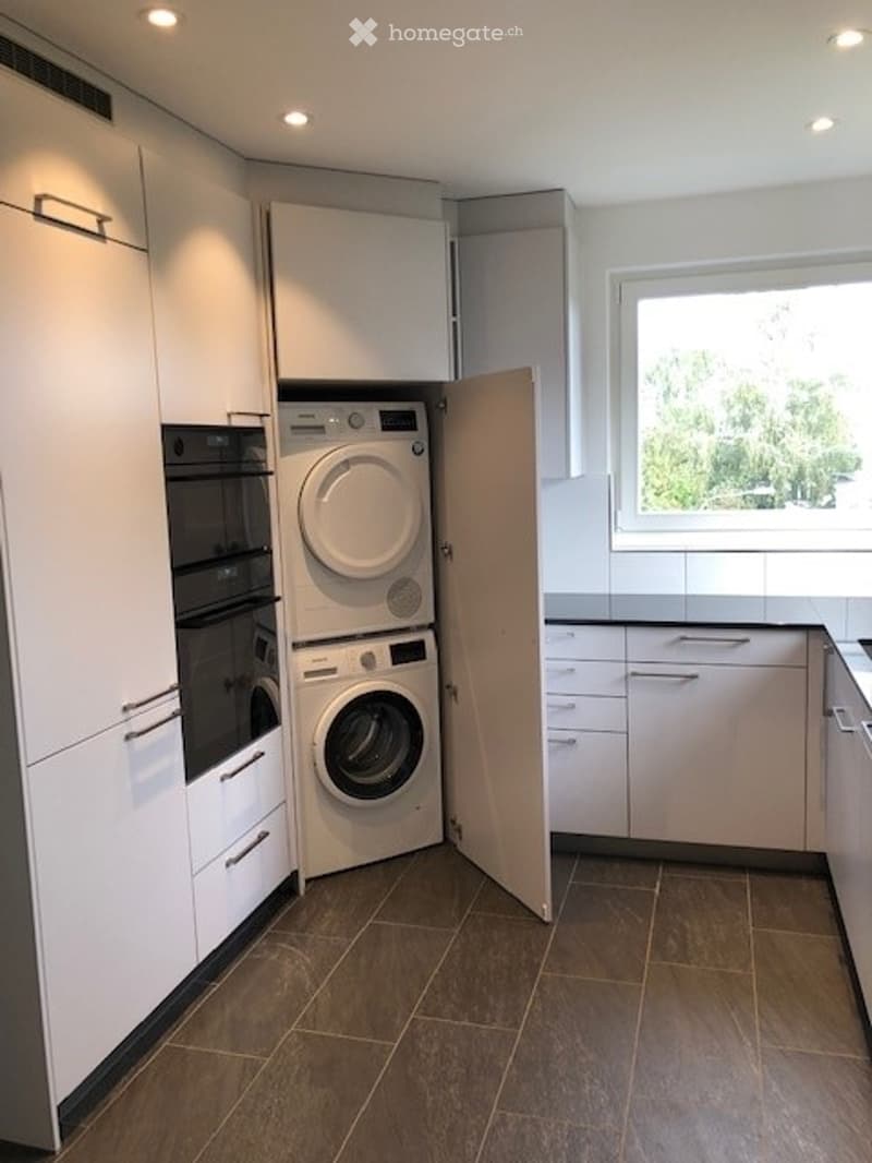 Modern kitchen with white cabinets, a built-in washing machine, and an oven. The space features a large window with a view of greenery outside.