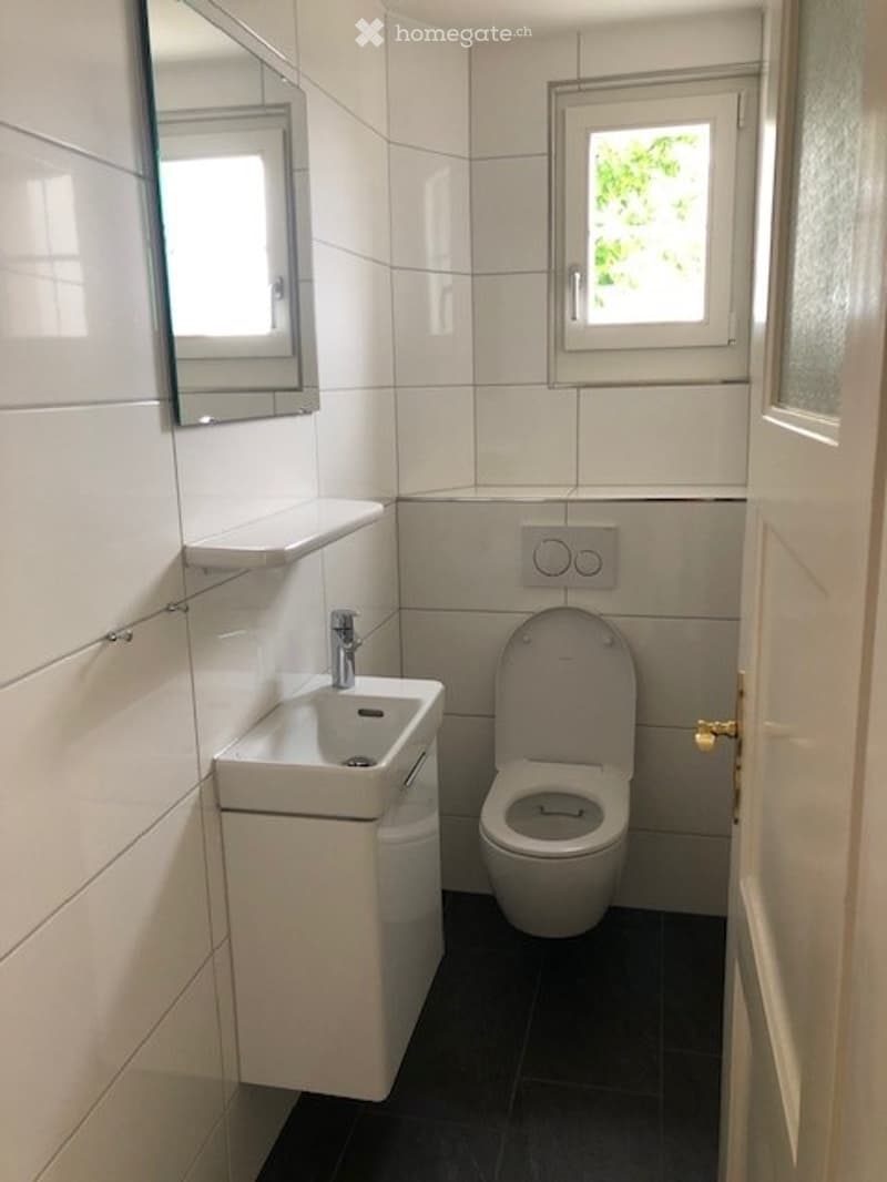 Modern bathroom with white tiles, a white sink, a toilet, a mirror, and a small window letting in natural light.