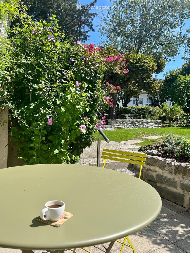 A small outdoor seating area with a green table, yellow chair, and a white coffee mug on a coaster, surrounded by lush flowering bushes and a well-maintained lawn.