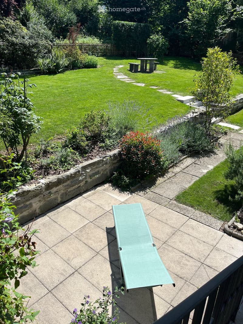 Well-maintained garden with a stone patio, sun lounger, small table and bench, surrounded by greenery and shrubs.