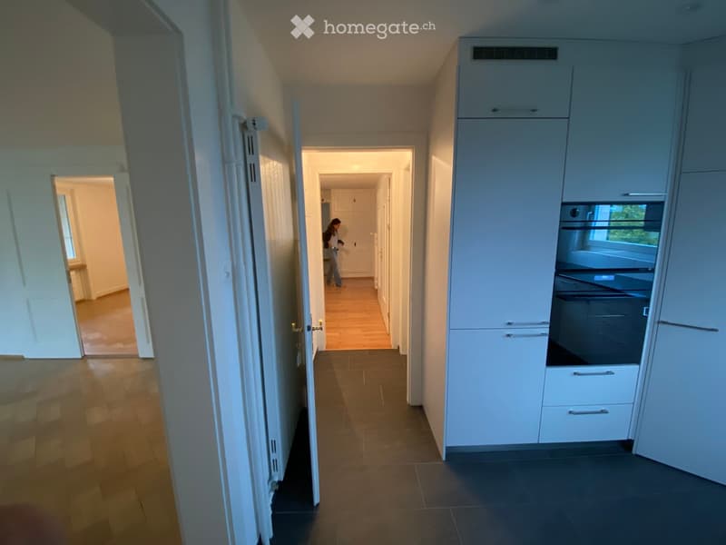 Empty hallway with wooden flooring, white walls, and multiple doors leading to other rooms. A modern kitchen with white cabinets and stainless steel appliances is visible on the right side.