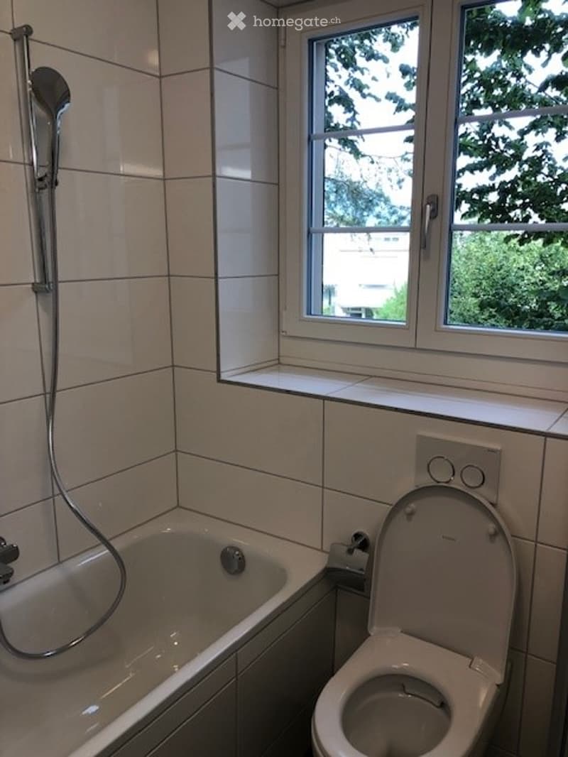 Bathroom with a bathtub, toilet, and showerhead, featuring white tiled walls and a window with a view of greenery.