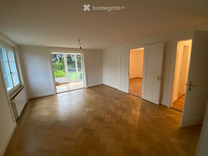 Empty room with wooden flooring, white walls, and large windows. Open doors lead to another room and a garden area.