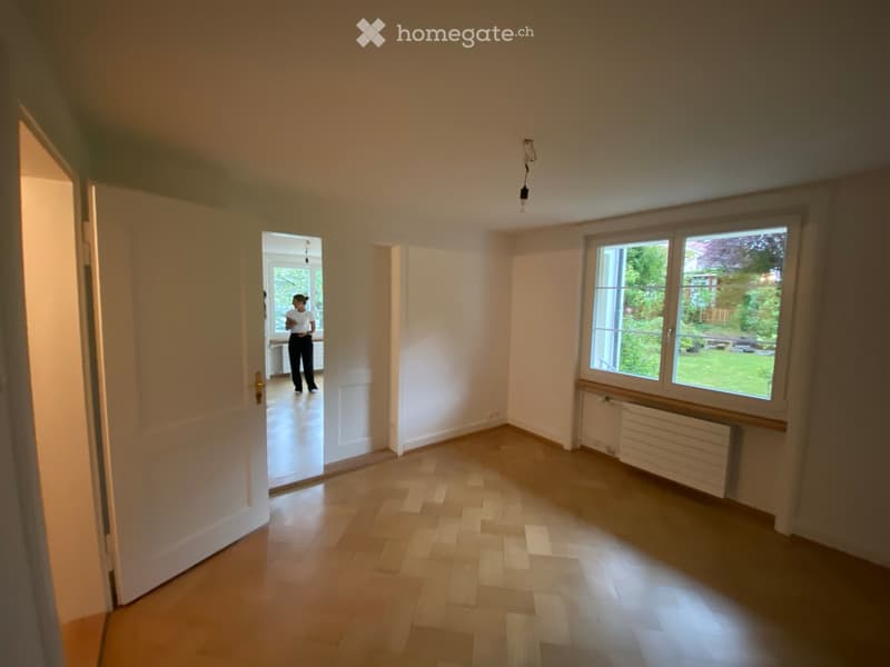 Empty room with wooden flooring, white walls, large windows, and a radiator. The room is unfurnished and has an open doorway leading to another space.