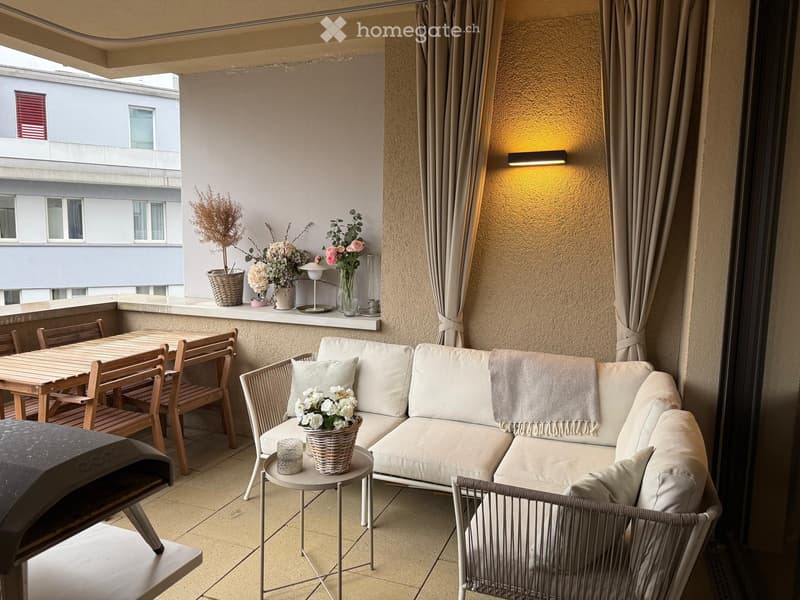 Balcony with a white sofa, small round table, wooden dining table, and chairs, decorated with flower pots and a wall-mounted light fixture.
