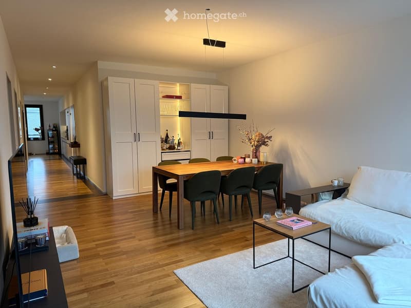 Modern, well-lit living and dining area with a white sofa, coffee table, dining table with chairs, and a small kitchen area visible in the background.