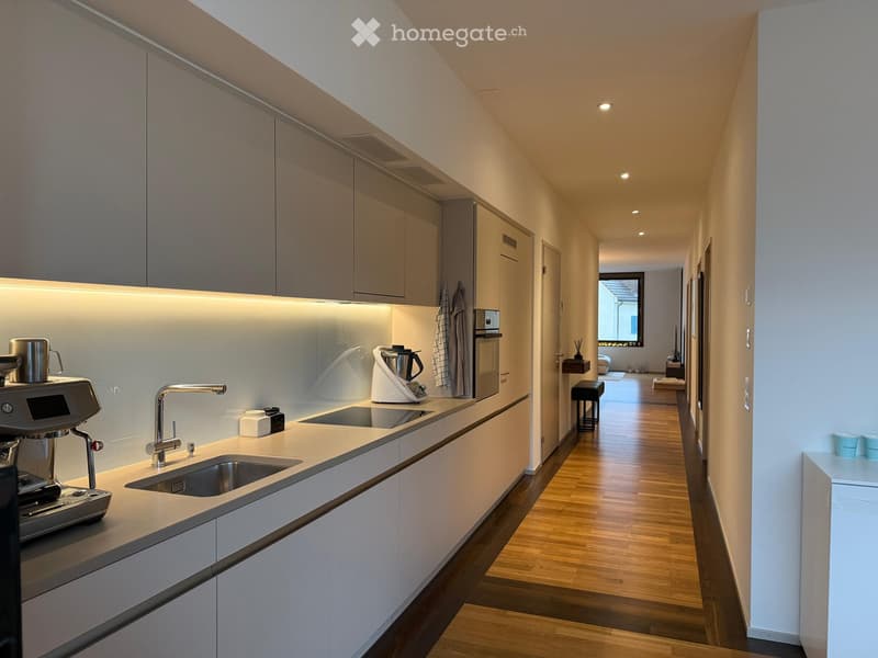 Modern kitchen with white cabinets, a stainless steel sink, a coffee machine, and a sleek faucet. The hallway leads to another room with wooden flooring.