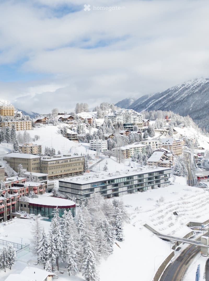 Aerial view of a snow-covered town with multiple buildings, surrounded by snow-covered trees and mountains.