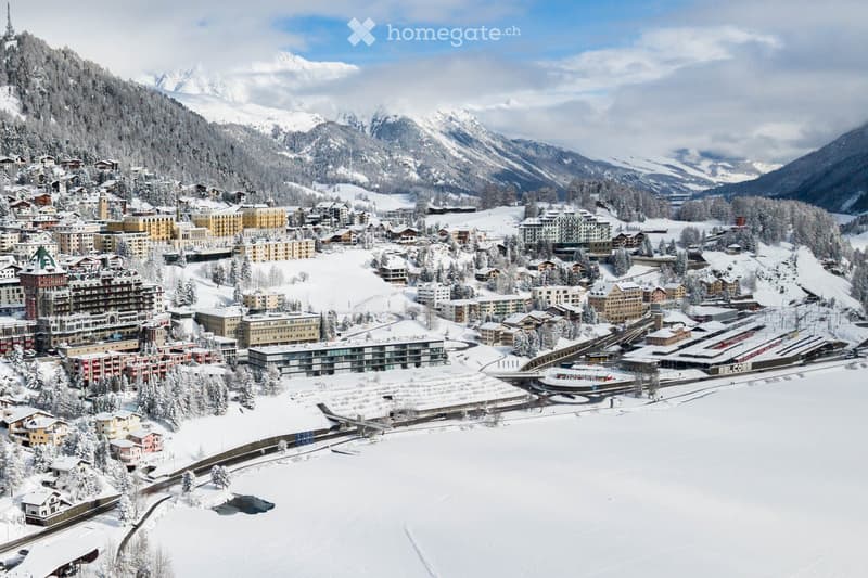 Aerial view of a snowy town nestled in a mountainous landscape, featuring numerous buildings, a ski slope, and a train station.