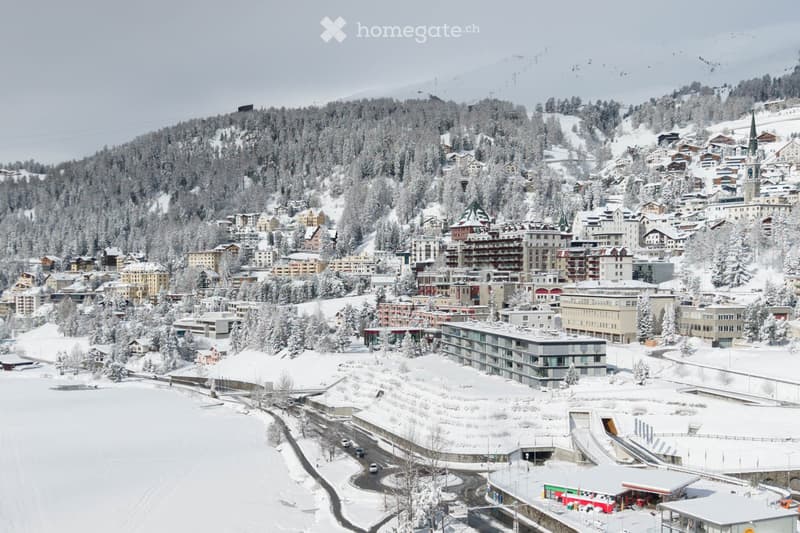 Aerial view of a snow-covered town nestled in a mountainous area, with numerous buildings, roads, and a church tower visible.