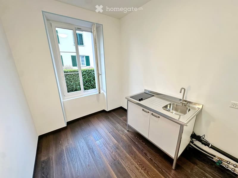 Modern kitchen with white cabinets, stainless steel sink, and stove, featuring wooden flooring and large window with view of greenery.