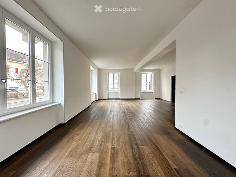 Empty room with wooden flooring, white walls, and large windows, featuring a slanted ceiling and multiple doorways.